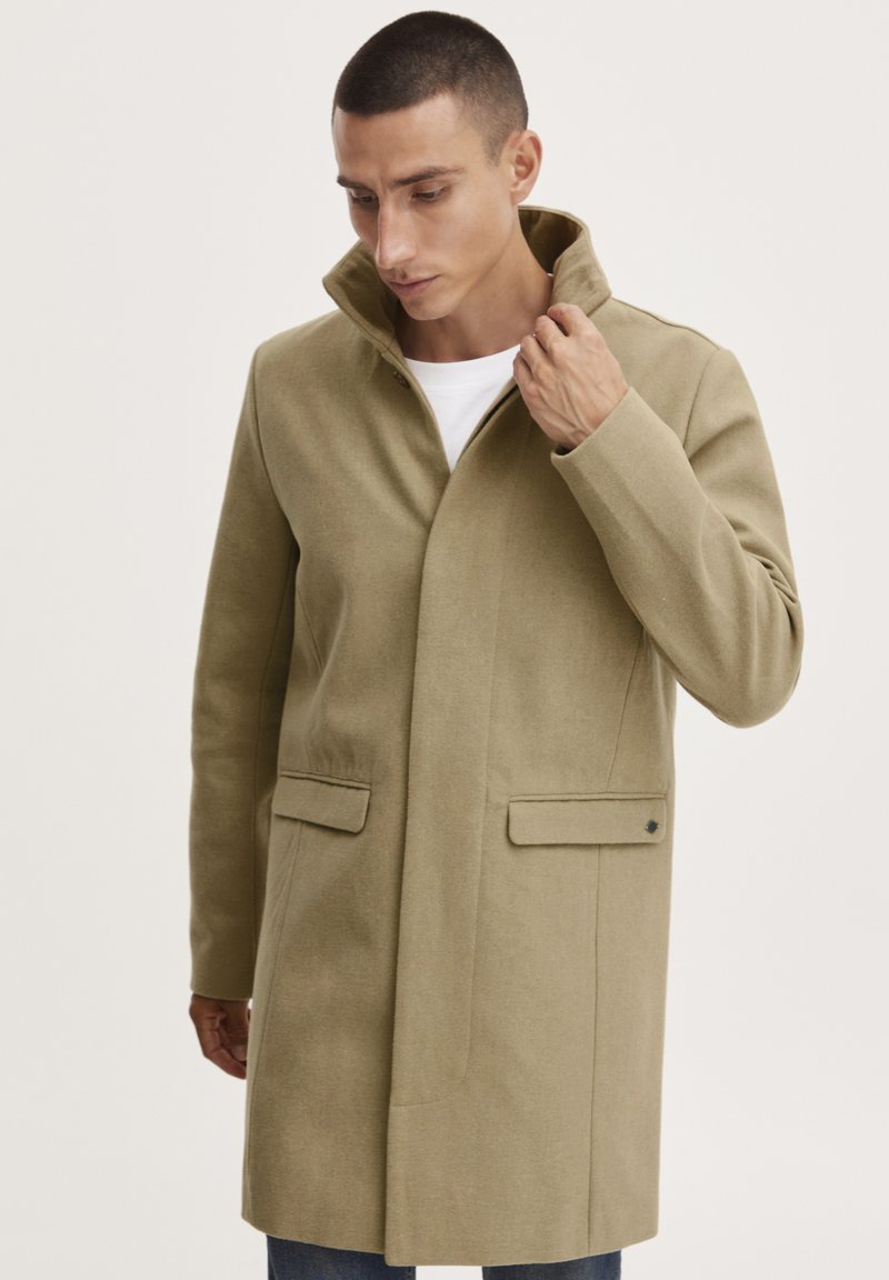 Cappotto di lana beige con colletto alto, due tasche frontali e vestibilità su misura, caratterizzato da una texture liscia e dettagli di design minimalisti.