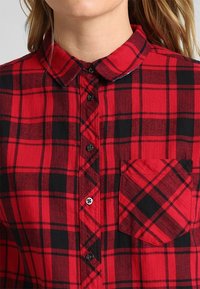 Camisa de franela a cuadros rojos y negros con cuello redondeado, botón al frente y un bolsillo en el pecho que presenta un patrón a juego. Textura suave.