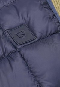 Mornarići kvadratasti materijal s teksturiranom logotipskom značkom angularnog dizajna. Materijal izgleda izdržljivo s suptilnim sjajem.