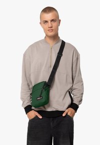 Grön crossbody-väska med en texturerad yta, dragsnodd och logotyp-patch, bärs över en ljusgrå, zippered sweatshirt och svarta byxor.