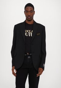 Blazer nero con una vestibilità sartoriale, indossato sopra una T-shirt nera con testo dorato, abbinato a jeans scuri. Dettaglio riconoscibile: etichetta sul blazer.