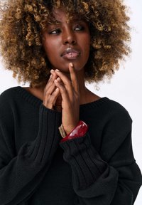 Pull en tricot noir avec un col large et des poignets côtelés. Le modèle porte un bracelet rouge épais et tient les mains ensemble, mettant en avant un teint naturel.