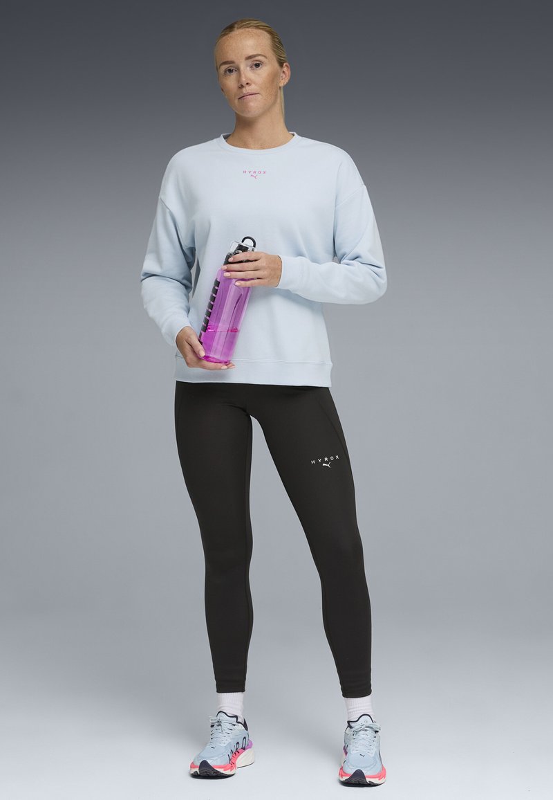 Femme en sweat-shirt bleu clair et leggings noirs tenant une bouteille d'eau rose, debout sur un fond dégradé gris.