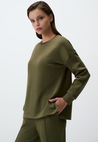 Felpa a maniche lunghe verde oliva con vestibilità ampia, scollatura rotonda, spacchi laterali e una texture liscia. Abbinata a pantaloni coordinati.