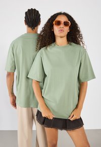Lys grønn bomull t-skjorte med korte ermer og rund hals, båret av en modell som står foran en annen person. Minimalistisk design.
