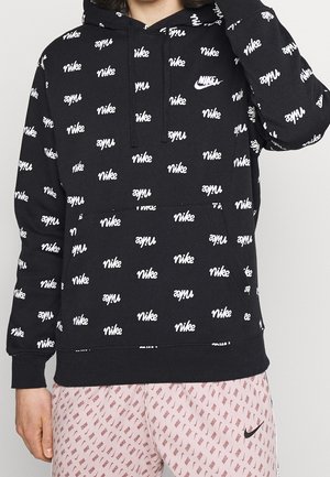 Osoba nosząca czarną bluzę z kapturem i różowe szorty, oba pokryte powtarzającymi się białymi logo Nike, z białym znakiem Nike na klatce piersiowej i szortach.