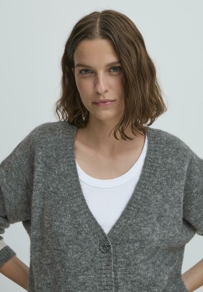 Šedý pletený kardigán s výstrihom do V, s jedným čiernym gombíkom, nosený preko bieleho topu. Mäkka textúra, voľný strih a spustené ramená.