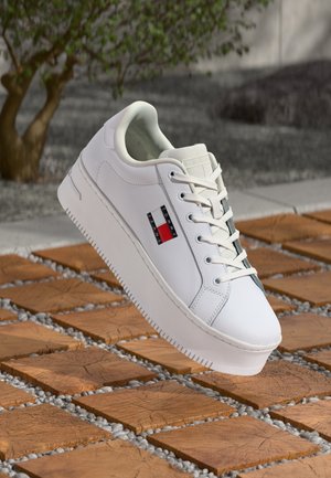 Valkoinen plattauskenkä, jossa Tommy Jeans -logo sivussa, valkoiset nauhat, esillä puisella laattalla ja sorapintaisella ulkoalueella.