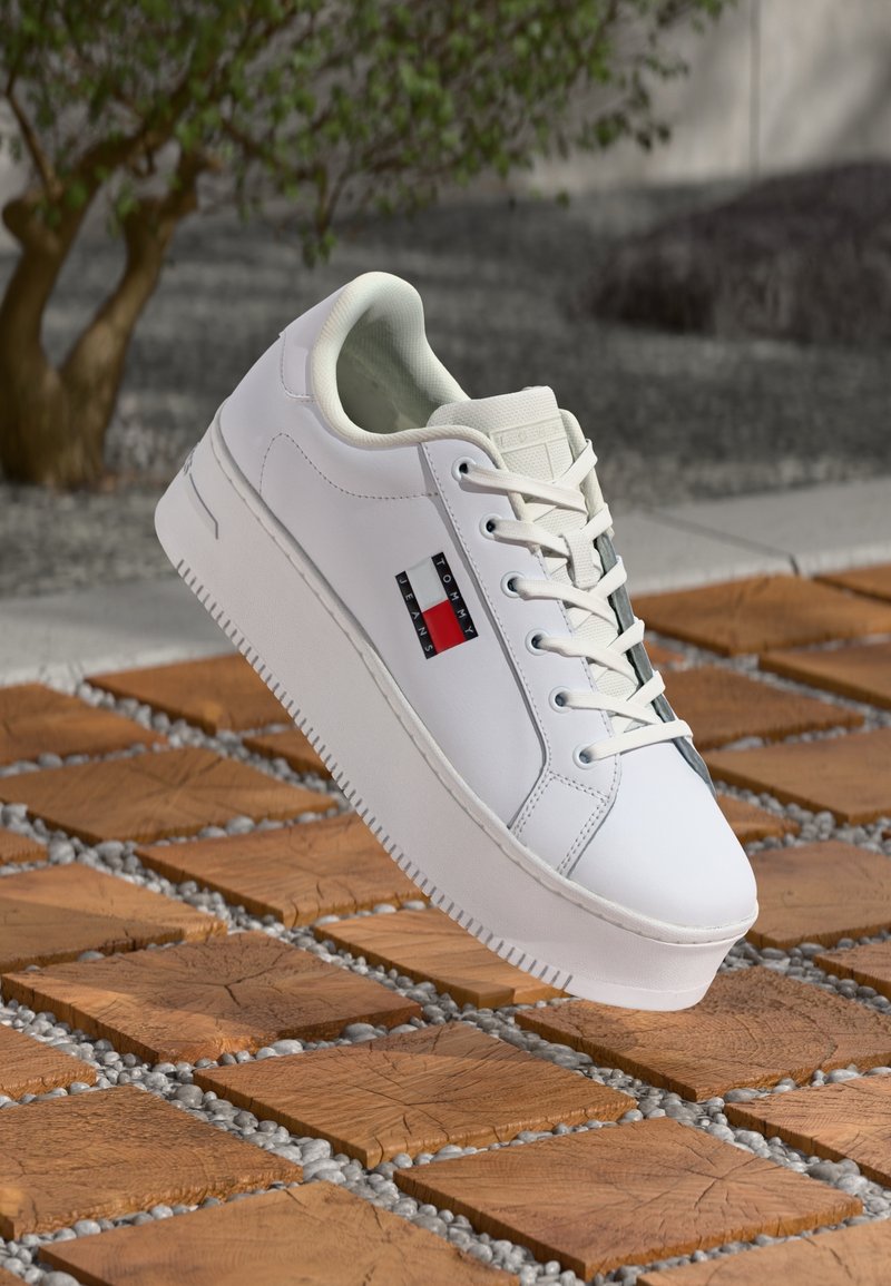 Baskets à plateforme blanches avec logo Tommy Jeans sur le côté, lacets blancs, présentées sur une dalle en bois et un sol de galets en extérieur.