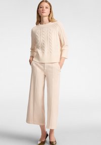 Crème kabelgebreide sweater met ¾ mouwen, gecombineerd met lichtbeige brede broek en olijfgroene hakken. Model staat met handen in de zakken.