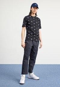 Camiseta negra con logotipos de Nike repetidos, combinada con pantalones oscuros y zapatillas Nike blancas. El modelo también lleva una gorra Nike azul.