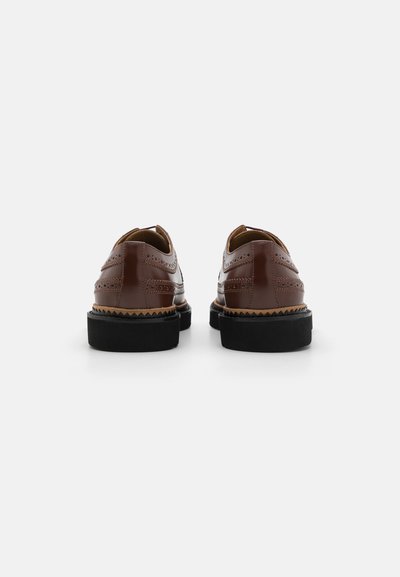 Brune lædersko med perforerede detaljer, kontrasterende sorte gummisulder og en rund tå. Simpelt design med minimale accenter.