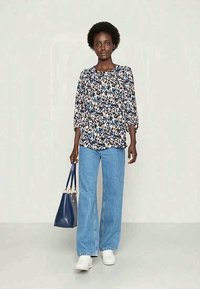 Blusa estampada com flores e mangas três quartos, combinada com jeans de perna larga azul claro e ténis brancos; bolsa azul escura com detalhes neutros.
