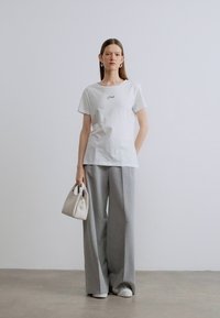 Λευκό βαμβακερό T-shirt με κοντά μανίκια και μικρό μαύρο λογότυπο, σε συνδυασμό με γκρι παντελόνι πλατιάς γραμμής και λευκή τσάντα.