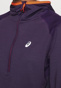 Sudadera deportiva morada con diseño de media cremallera, acentos naranjas en el cuello y detalles de canalé texturizados en los hombros. Presenta un logo.