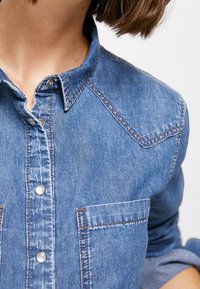 Camicia in denim di colore azzurro chiaro con colletto, due tasche sul petto e dettagli di cucitura ben visibili. Realizzata in morbido denim texturizzato.