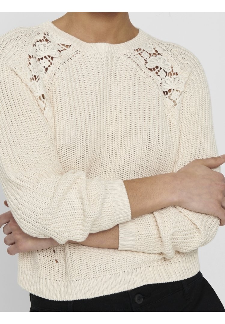 Pull tricoté écru avec des découpes en dentelle florale sur les épaules, porté avec un pantalon noir, bras croisés sur le torse.