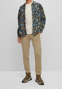 Man står med beiga byxor, bruna vandringsskor, vit skjorta och en blå, brun och vit mönstrad skjorta med knappar.