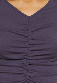 Detailní záběr na fialový top z řasené látky s výstřihem do V, který nosí osoba.