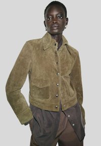 Veste en Daim vert olive avec un col, des boutons à pression et une poche frontale. Présente une coupe courte et une texture douce.