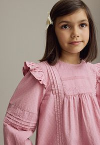 Lyserød bomuldsdragt med flæser ved ærmerne, lodrette folder og delikate broderede detaljer. Glat tekstur med små blomstermønstre.