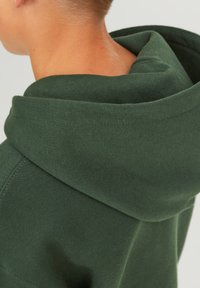 Sudadera con capucha de color verde oscuro y textura suave, que presenta una capucha redondeada y costuras reforzadas. La tela parece gruesa y cálida.