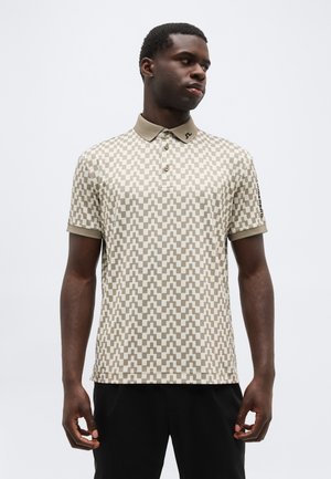 Mann trägt ein beige-weißes Polo-Shirt mit geometrischem Muster und kurzen Ärmeln sowie schwarze Hose, stehend vor einem schlichten weißen Hintergrund.