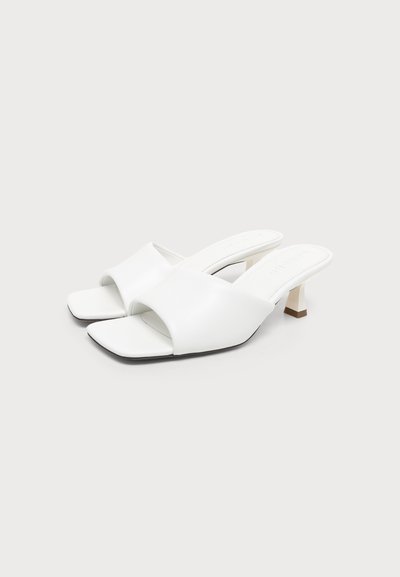 Paire de sandales blanches à bout ouvert avec petits talons blocs et large bride sur le dessus, sur fond gris clair.