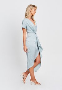 Svetlo modra satenasta obleka z V-izrezom, kratkimi rokavi in asimetrično robom. V kombinaciji z belimi sandali z odprtim prstom. Gladka tekstura.