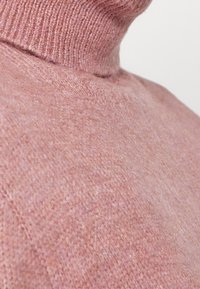 Maglione a collo alto rosa chiaro con una maglia morbida e texturizzata. La vista ravvicinata mostra il colletto a coste e le fibre sottili. Design semplice e aderente.