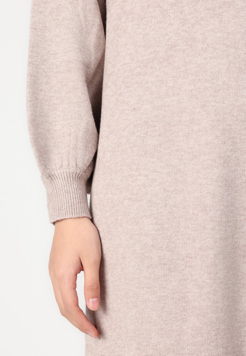 Jasnoróżowy sweter z dzianiny z prążkowanymi mankietami. Miękka tekstura i luźny krój. Widoczna dłoń umieszczona blisko krawędzi. Minimalistyczny design.