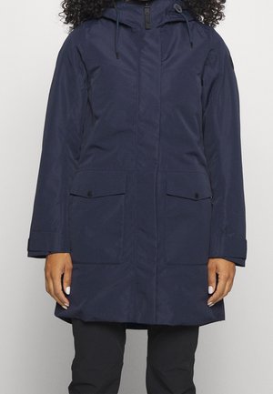 Kvinde iført en marineblå hættetrøje med forlommer og sorte bukser mod en ensfarvet lys baggrund.