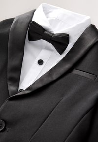 Veste de smoking noire à revers en satin portée sur une chemise blanche boutonnée de noir, assortie à un nœud papillon noir.