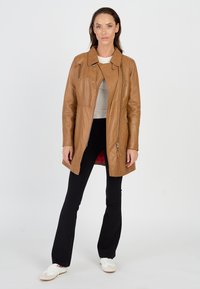 Beige Leder-Trenchcoat mit Schalkragen, Frontreißverschlüssen und langen Ärmeln. Getragen über einem hellen Oberteil und kombiniert mit schwarzen Hosen und weißen Schuhen.