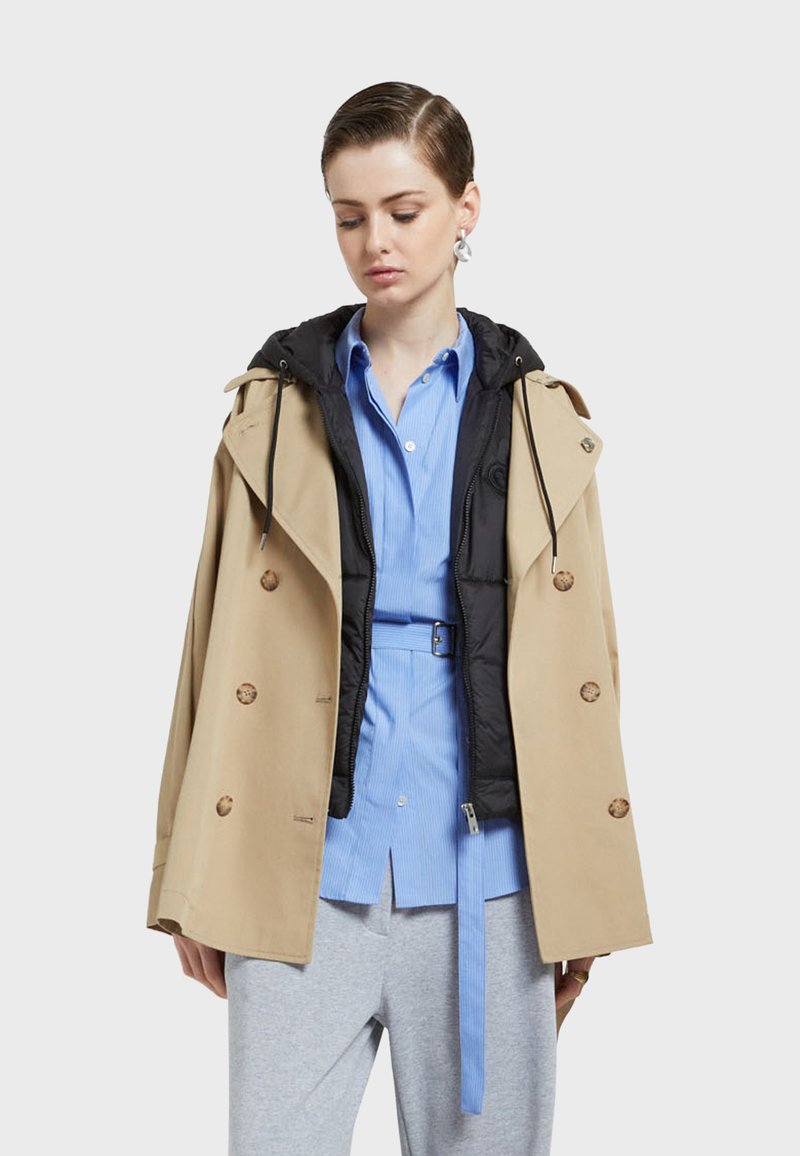 Beige trench coat with large brown buttons, layered over a black zip-up jacket, paired with a light blue button-up shirt and gray pants.