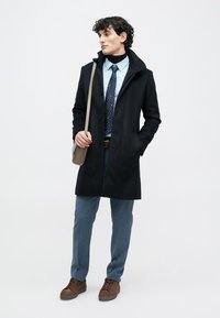 Uomo che indossa un cappotto blu navale, una camicia azzurro chiaro, una cravatta a fantasia, pantaloni blu, scarpe marroni e che porta una borsa a tracolla beige, in piedi contro uno sfondo bianco.