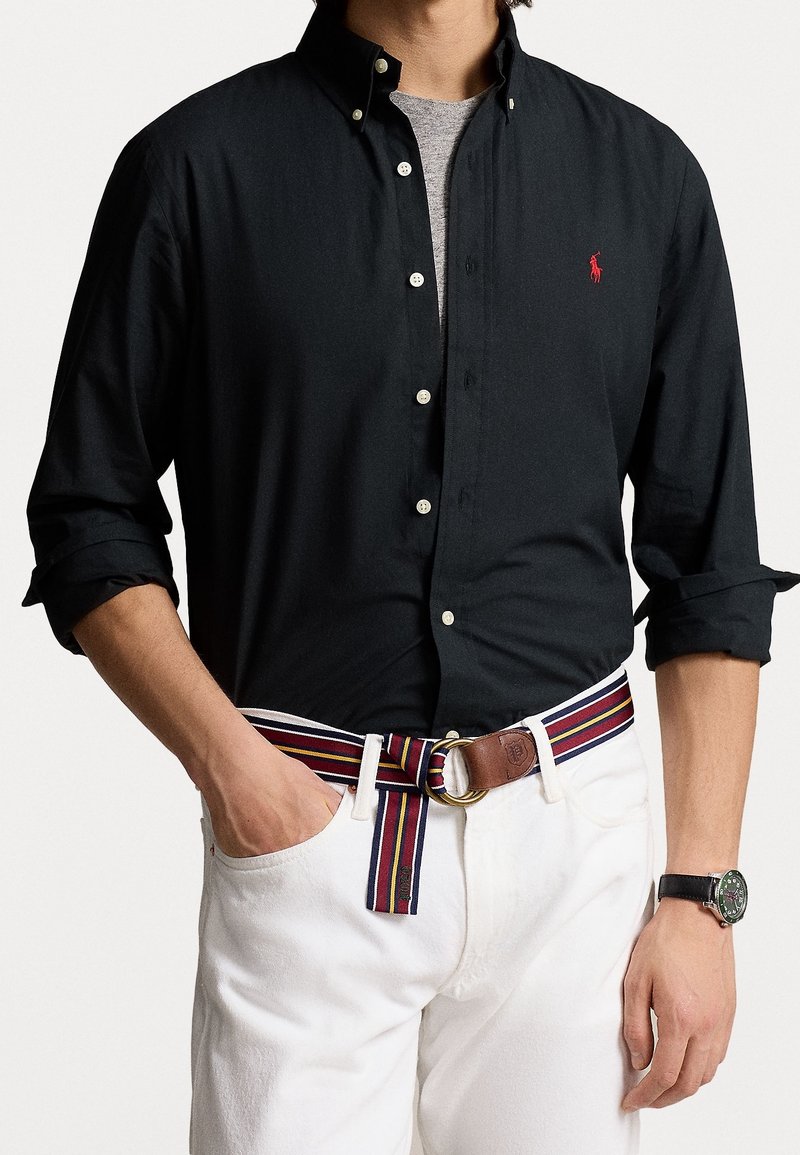 Svart skjorta med knappar och krage, vita denimbyxor, randigt bälte med läderdetaljer och en klocka med rund urtavla.