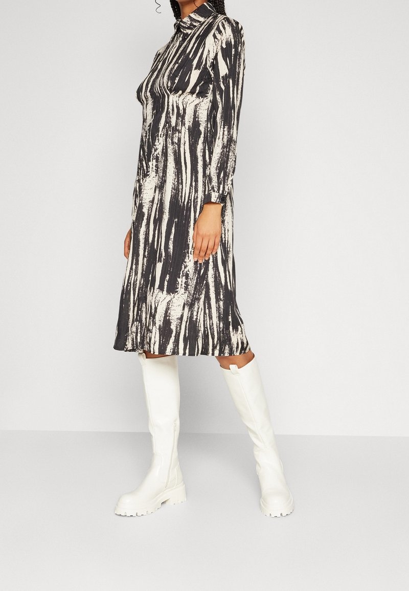 Femme portant une robe midi à manches longues avec un imprimé abstrait noir et blanc et des bottes blanches épaisses jusqu'au genou, debout de profil.
