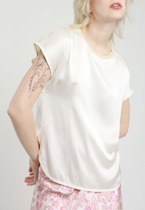 Persona che indossa una maglietta a maniche corte in raso color crema e pantaloni rosa con motivo paisley, mostrando un tatuaggio sul braccio superiore sinistro.