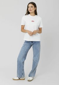 T-shirt en coton blanc à manches courtes, avec un texte rouge brodé sur la poitrine. Associé à un jean en denim bleu clair et des baskets blanches.