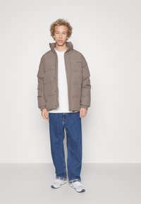 Brown padded jacket with a high collar, paired with a white sweater and blue loose jeans. Notable details include snap buttons and logo.