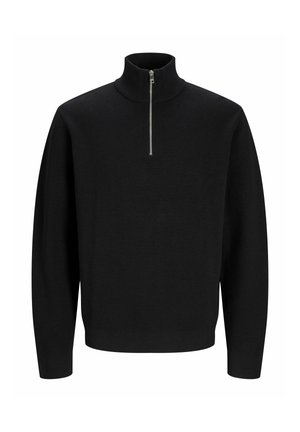 Jeune personne aux cheveux bouclés portant un pull noir à quart de zip, une chemise à carreaux, un large jean bleu et des chaussures beiges, regardant sur le côté.
