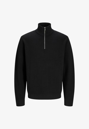 Ung person med krøllet hår iført en sort kvart-zip sweater, ternet skjorte, brede blå jeans og beige sko, kigger til siden.