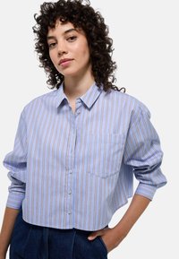 Chemise à boutons cropped rayée bleue et blanche avec une poche. Présente une coupe décontractée et des manches longues avec des poignets à boutons.