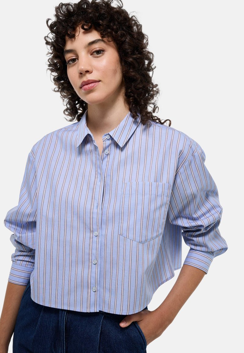 Chemise à boutons cropped rayée bleue et blanche avec une poche. Présente une coupe décontractée et des manches longues avec des poignets à boutons.