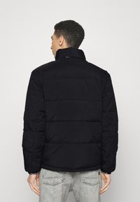 Giacca a vento nera con colletto alto, texture trapuntata e polsini elasticizzati, indossata sopra jeans grigi. Il design presenta un taglio dritto sul retro.