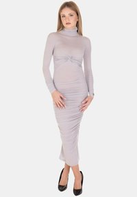 Abito grigio a maniche lunghe con collo alto, dettagli arricciati in vita e una silhouette aderente, abbinato a tacchi neri.