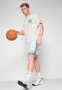 Svijetlosmeđi majica kratkih rukava s zelenom grafikom na prsima, uparen s bijelim šorcima sa šarenim cvjetnim rubom i crnim Nike logom.