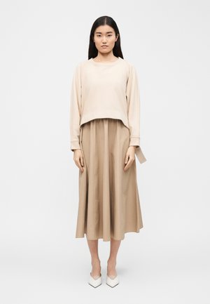 Donna con capelli lunghi neri che indossa una maglia beige a maniche lunghe, una gonna midi abbinata e décolleté bianche con punta appuntita, in piedi davanti a uno sfondo semplice.