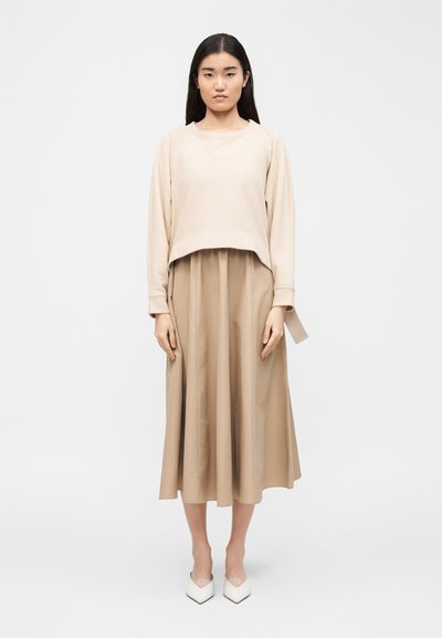 Femme aux longs cheveux noirs portant un haut beige à manches longues, une jupe midi assortie et des talons blancs à bout pointu, debout devant un fond uni.